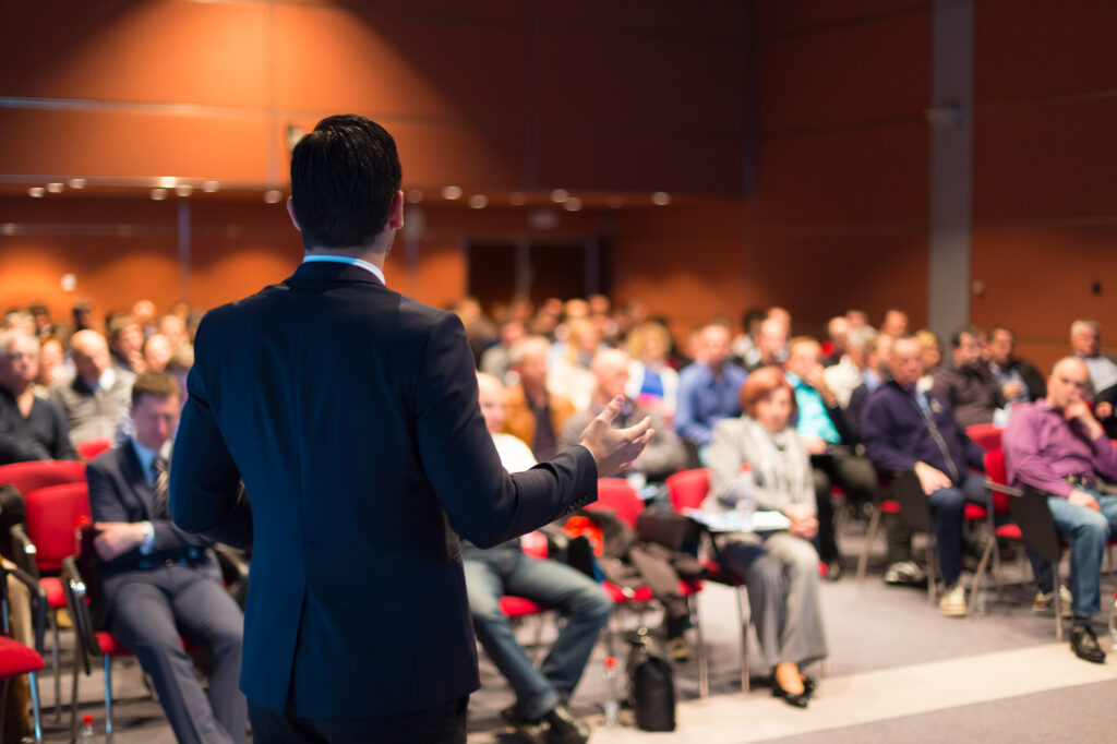 Speaker presenting to a large audience in conference room.